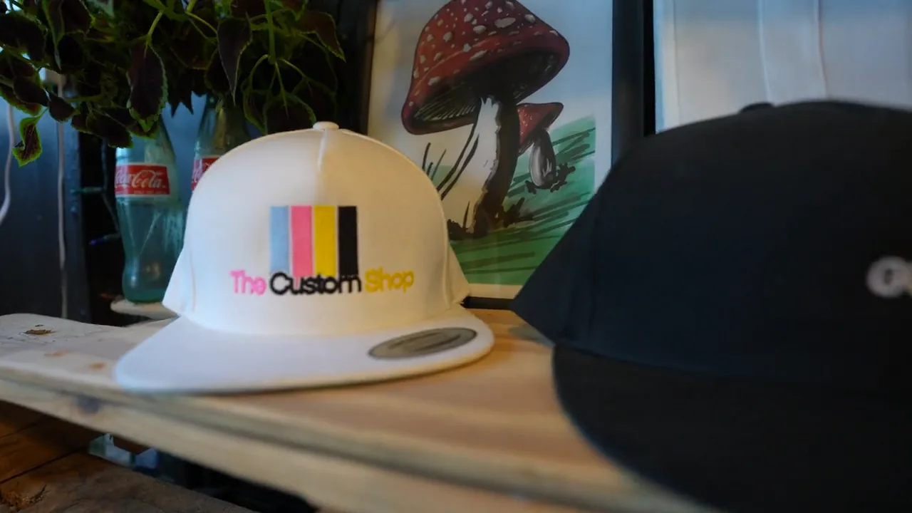 A gentle handheld move settles on a white Custom Shop cap, with colorful branding and artwork behind it.