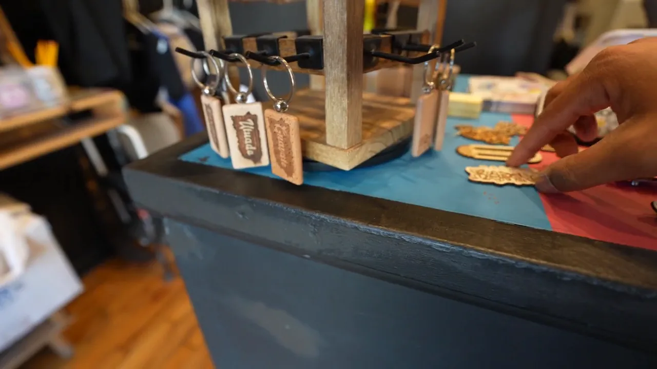 A hand arranges cut wooden pieces beside engraved keychains on a red and blue counter.