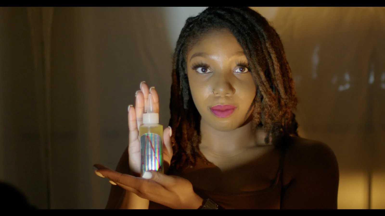 A woman holding lip gloss beside her face in golden light.