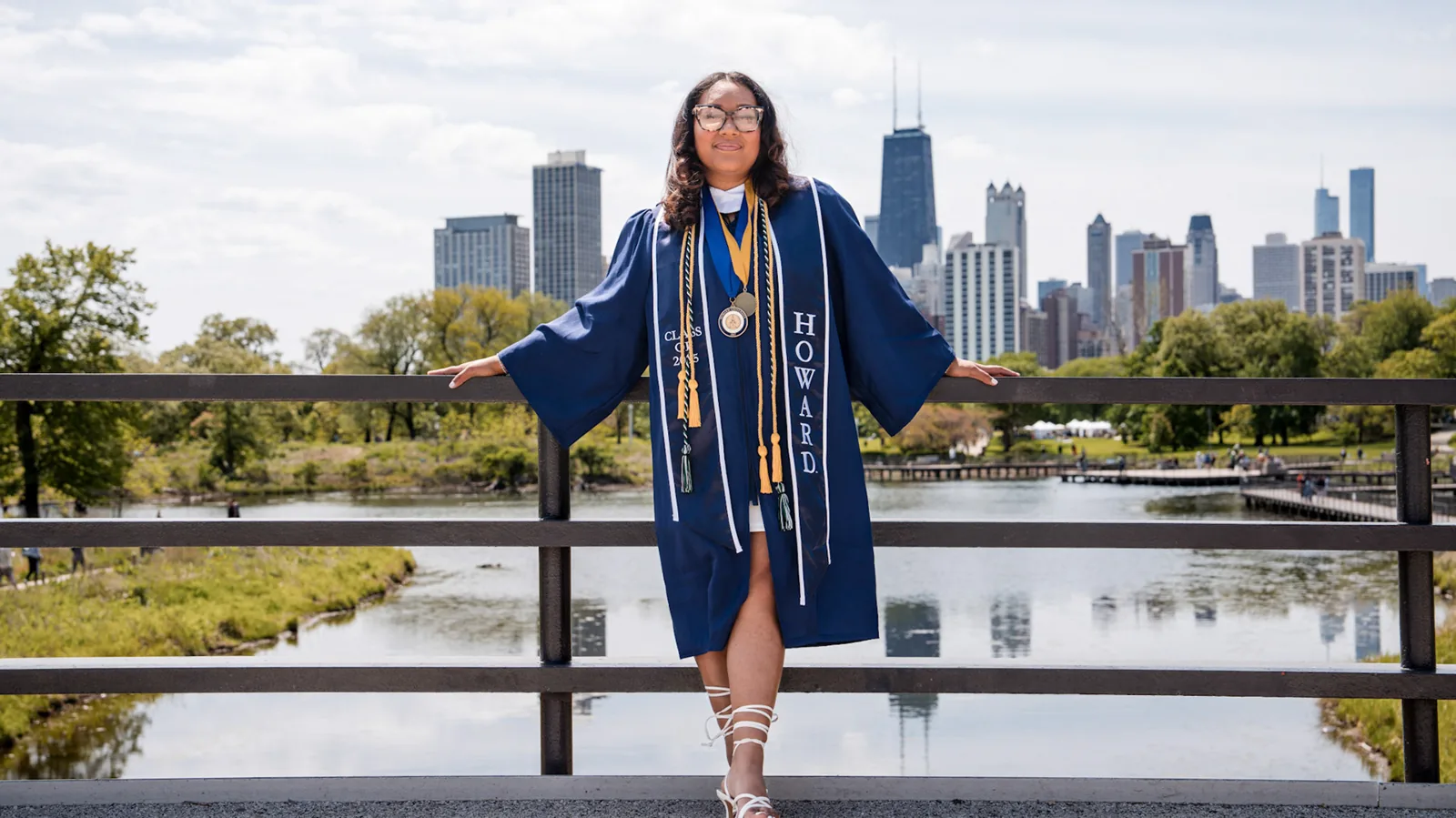 A graduate in a blue gown beside the Chicago skyline.
