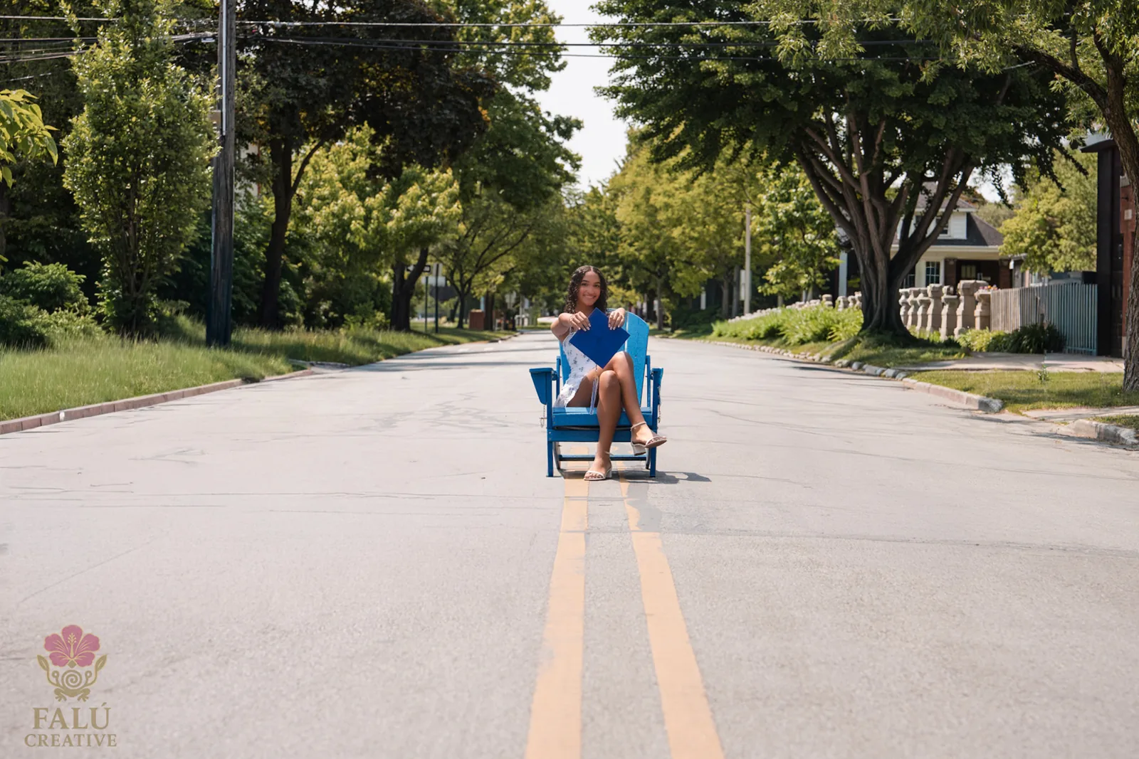 A graduate seated in the center of an empty tree-lined street.