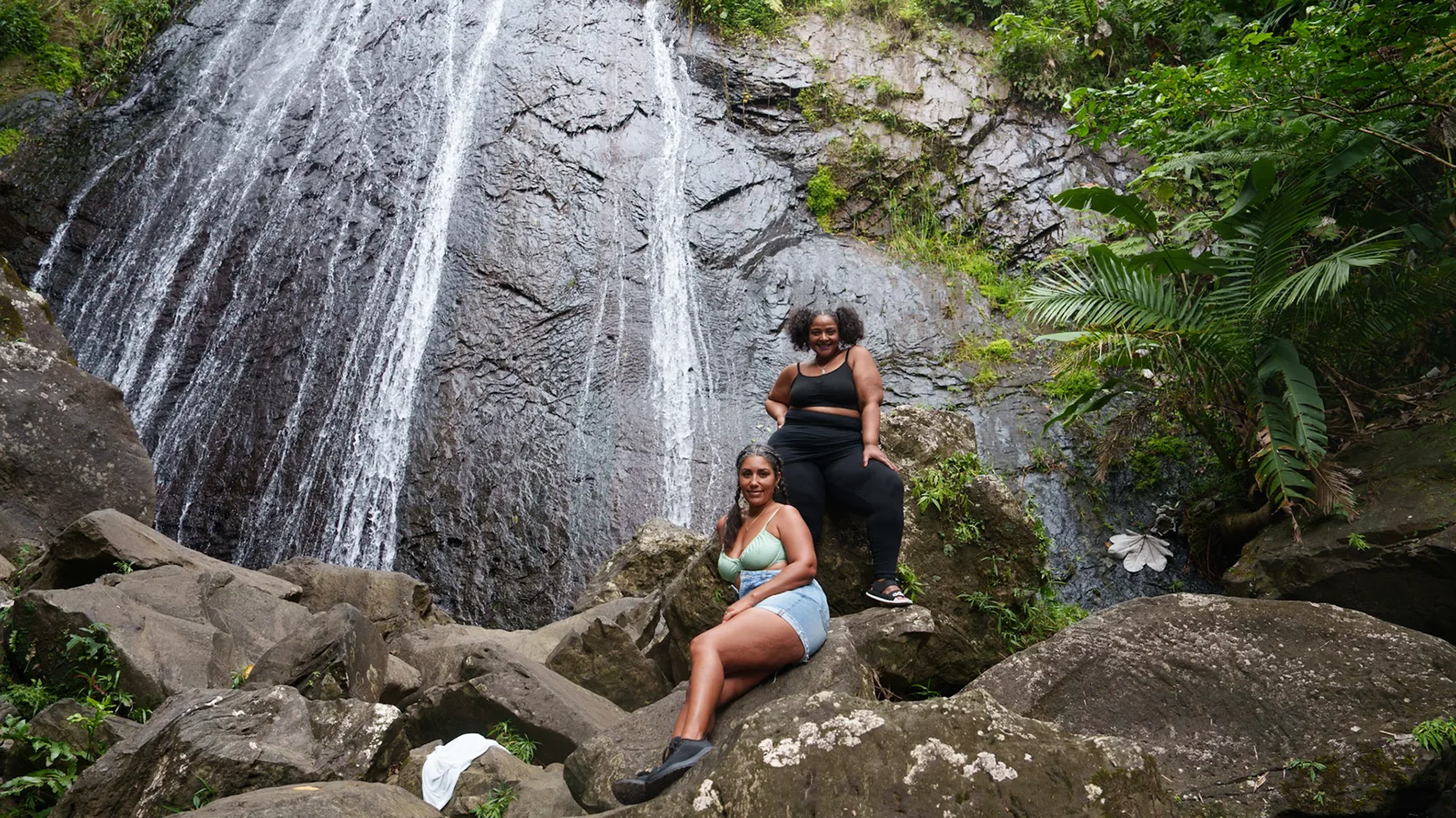 Two women sitting on rocks beneath a tall waterfall.