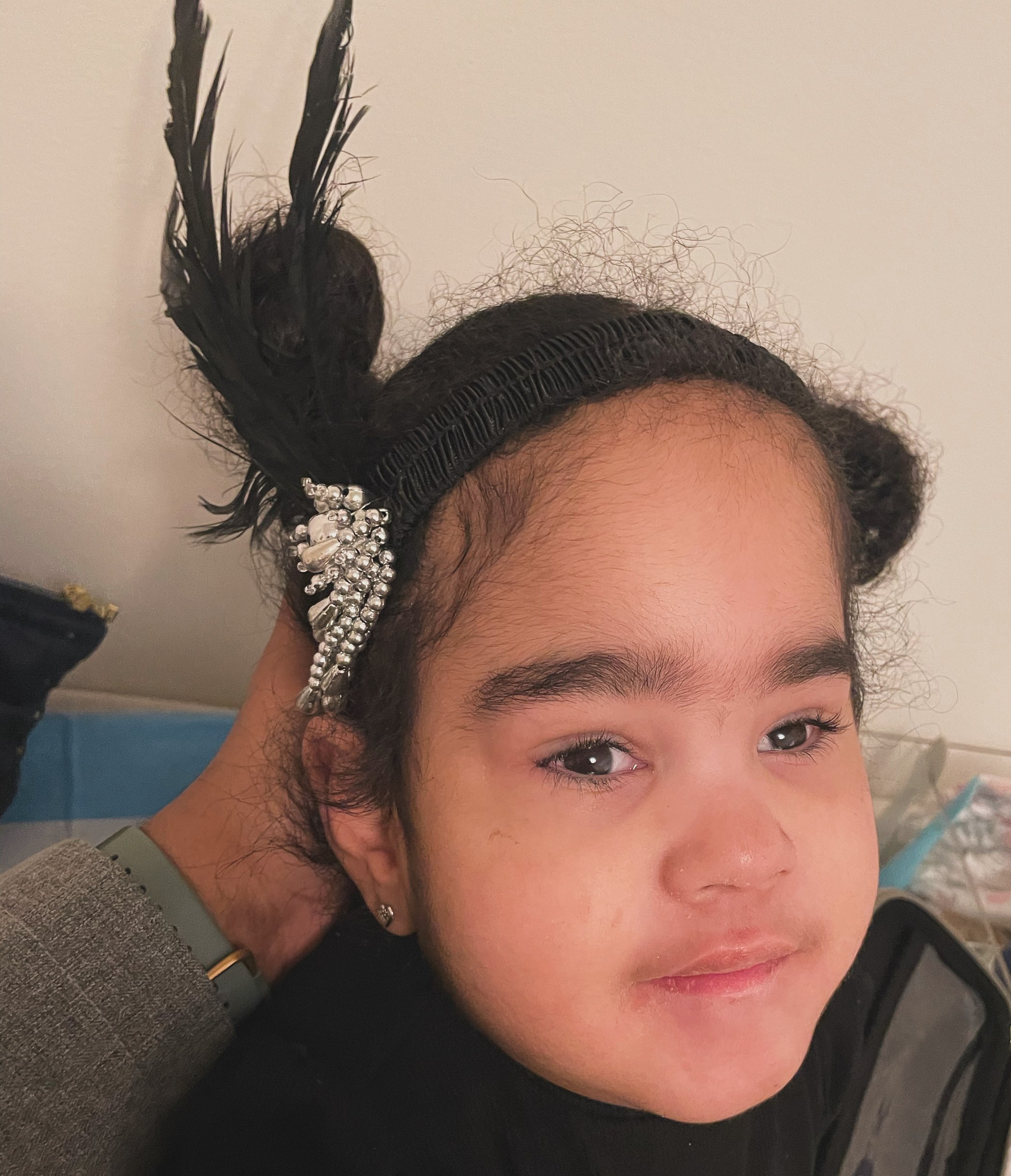 Havana, dressed up, looking straight at the camera. Her hair is up with a black feathered clip and a hand rests behind her head.