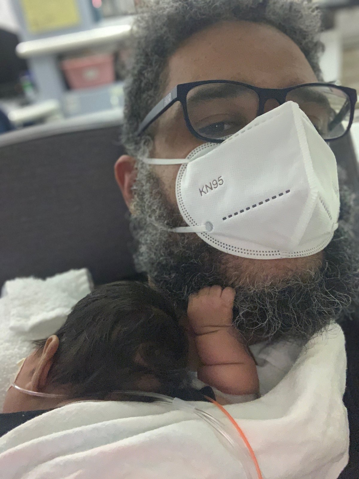Falú in a mask, holding newborn Havana against his chest in a hospital room.