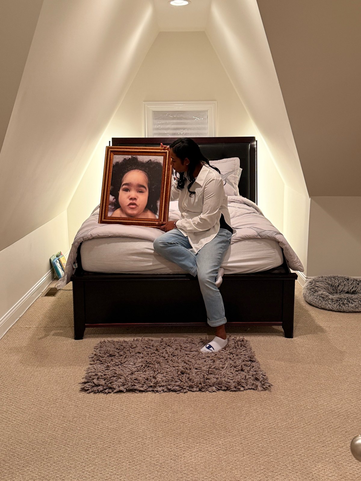 Indira sitting on a bed in an attic bedroom, holding a large framed portrait of Havana.