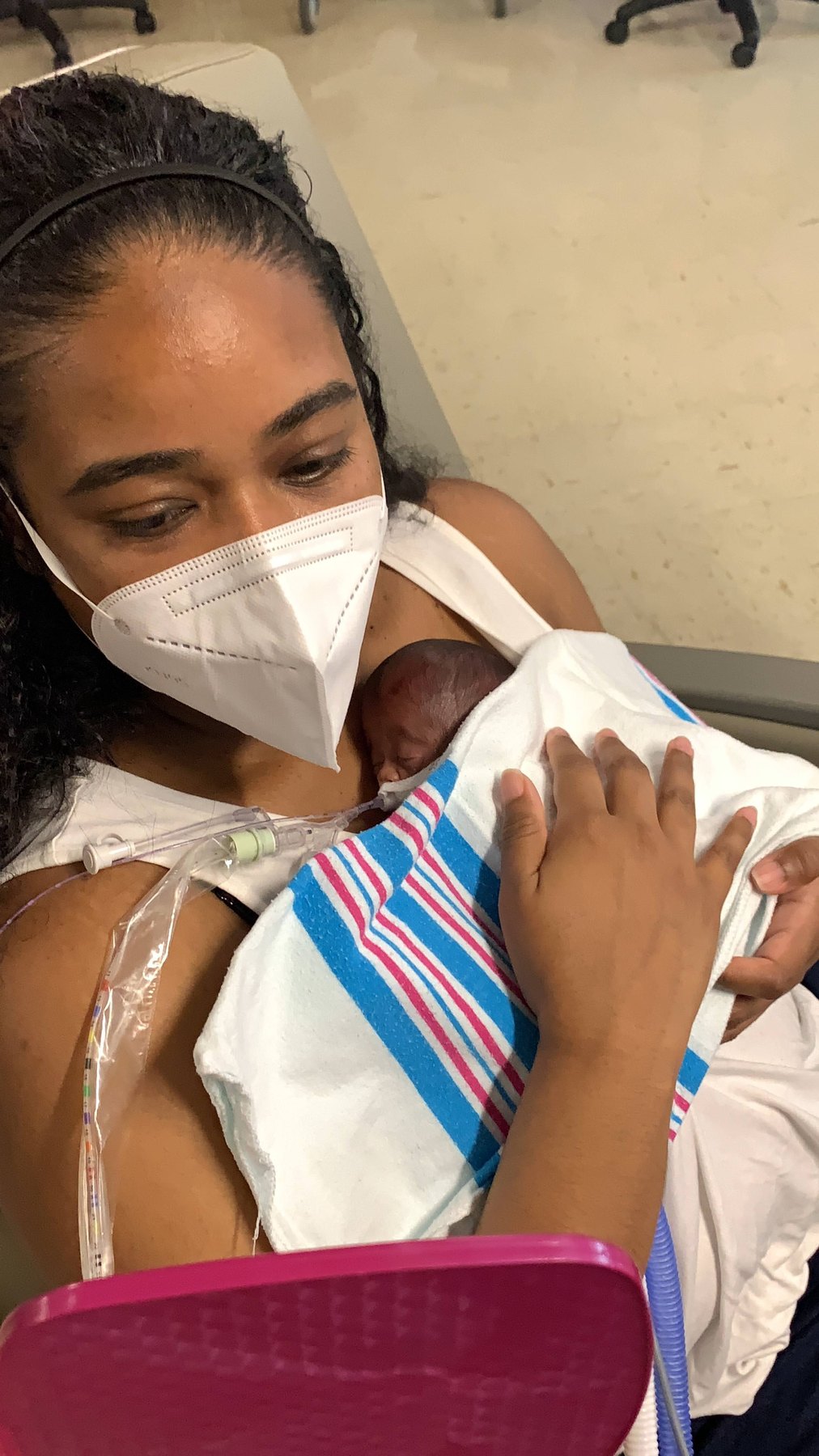 Indira, in a mask, looking down at newborn Havana on her chest, wrapped in a hospital receiving blanket.